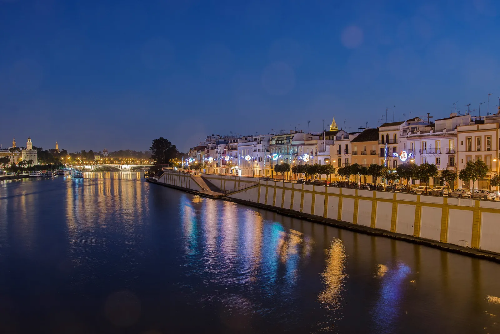 Guadalquivir v noci s osvětlenými domy na Calle Betis odrážejícími se ve vodě a siluetou Sevilly v dálce