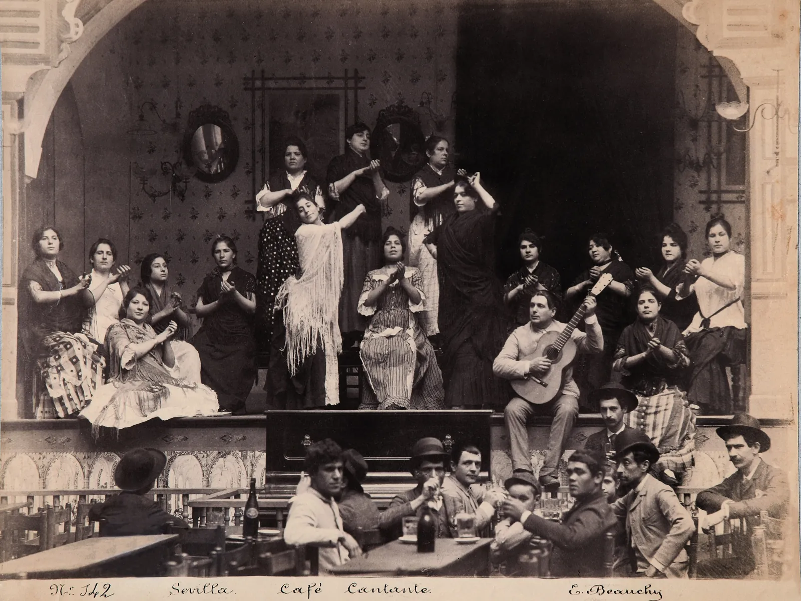 Stará černobílá fotografie café cantante v Seville, na níž skupina žen v šálách a mužů s kytarami pózuje na malém pódiu