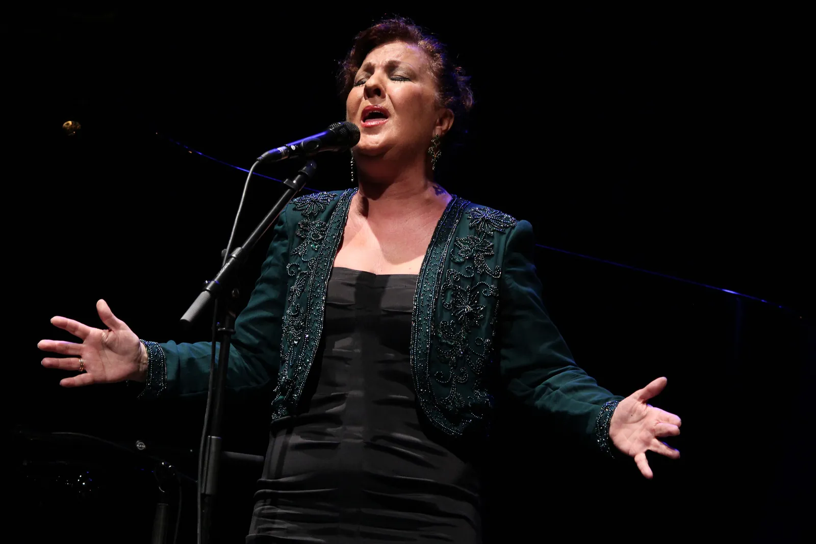 Flamenková zpěvačka na pódiu s otevřenou náručí a mikrofonem před sebou, osvětlená proti tmavému pozadí