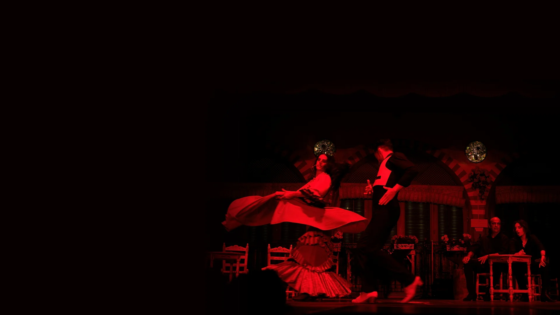 Dva tanečníci flamenca, žena v červených šatech uprostřed obratu a muž v černé vestě, na pódiu osvětleném do červena, se dvěma účinkujícími sedícími u malého stolku vpravo