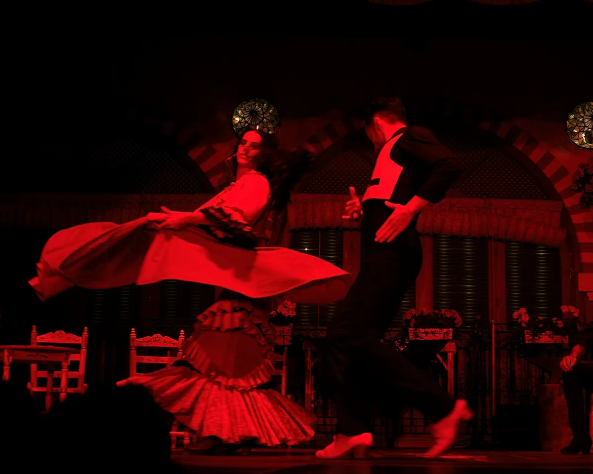 Flamenková tanečnice v červených šatech a tanečník v černém na jevišti tablao osvětleném do červena, dva účinkující sedící u stolku vpravo